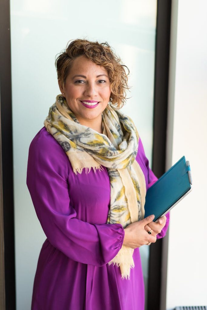 woman-smiling-and-holding-teal-book-1181424 Smiling woman in a purple dress and scarf holding a tablet indoors.